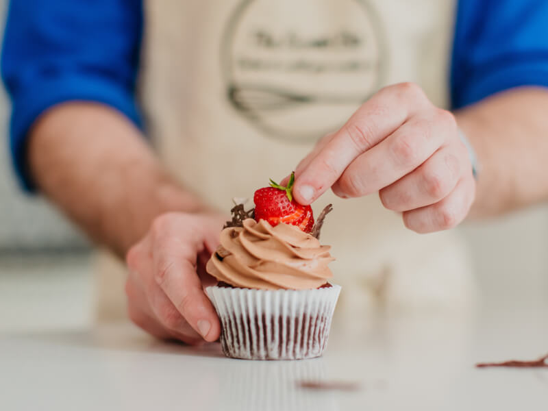 A man puts a strawberry on top of a chocolate butter iced cupcake as a finishing touch
